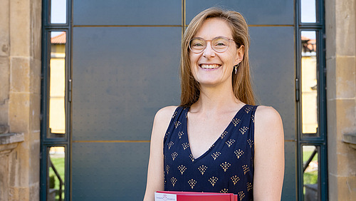 Eine junge Frau in einem eleganten Sommerkleid steht vor dem Eingang eines historischen Gebäudes. Sie lacht in die Kamera., in der Hand hält sie ine rote Mappe.