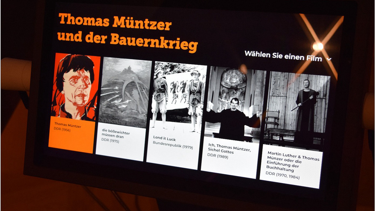 Das Bild zeigt eine digitale Infotafel in einem Museum. In orangener Schrift steht "Thomas Müntzer und der Bauernkrieg" geschrieben und darunter sind Filmausschnitte zur DDR.