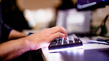 Nahaufnahme einer Hand, die auf einer beleuchteten Tastatur tippt. Im Hintergrund sind unscharf Computerbildschirme zu sehen, die durch das gedämpfte Licht beleuchtet werden.
