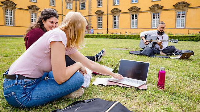 Three students are sitting together in the castle courtyard. One of them is playing the guitar.