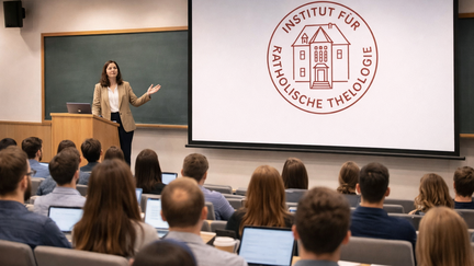 Eine Dozentin steht vorne in einem modernen H&ouml;rsaal an einem Pult und spricht zu einer voll besetzten Studierendengruppe. Auf einer Projektionsleinwand neben ihr ist das Logo des Instituts f&uuml;r Katholische Theologie zu sehen. Viele Studierende sitzen mit ge&ouml;ffneten Laptops in ansteigenden Sitzreihen.