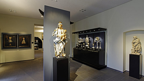 Ein Innenraum mit mehreren Ausstellungsst&uuml;cken. In der Mitte steht eine S&auml;ule oder Vitrine mit einem Objekt, das von Licht beleuchtet wird.  An den W&auml;nden h&auml;ngen Bilder oder Tafeln in gleichm&auml;&szlig;igen Abst&auml;nden. Rechts stehen dunkle Vitrinen mit weiteren Objekten.  Der Raum ist hell und hat einen glatten Boden.