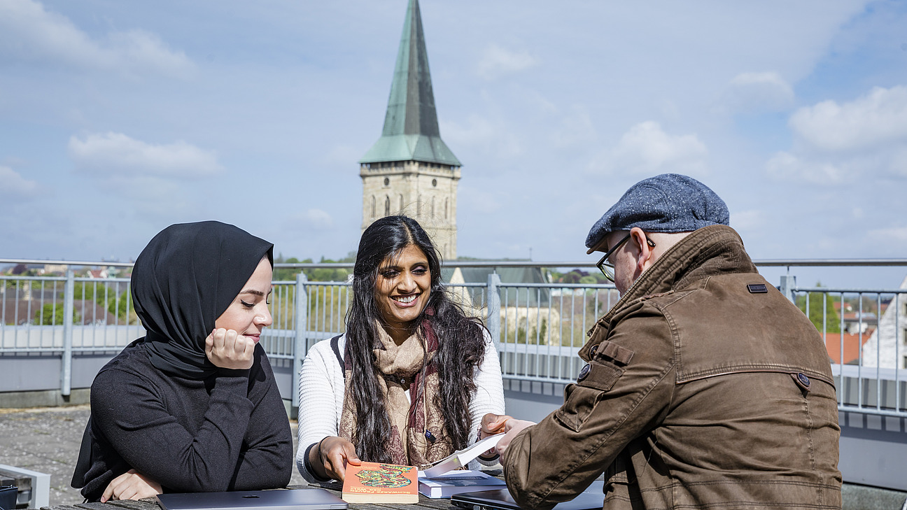 Drei Personen sitzen an einem Tisch im Freien. Eine von ihnen tr&auml;gt ein Kopftuch. Vor ihnen liegen mehrere B&uuml;cher, eins hat den Titel "Making the Arab World". Im Hintergrund ist die Sankt Katharinen Kriche zu sehen.
