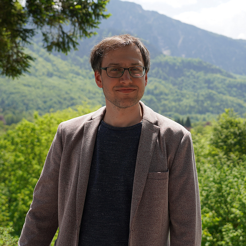 Ein Mann mit Brille steht vor einer Berglandschaft. Er trägt einen grauen Blazer und ein dunkles Oberteil.