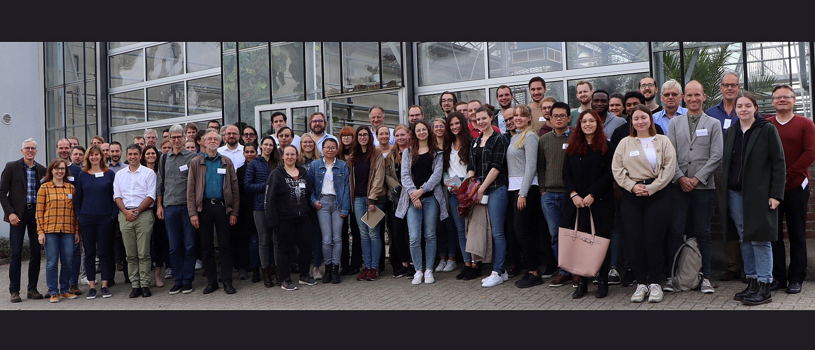 Eine sehr gro&szlig;e Personengruppe steht vor einem Gew&auml;chshaus