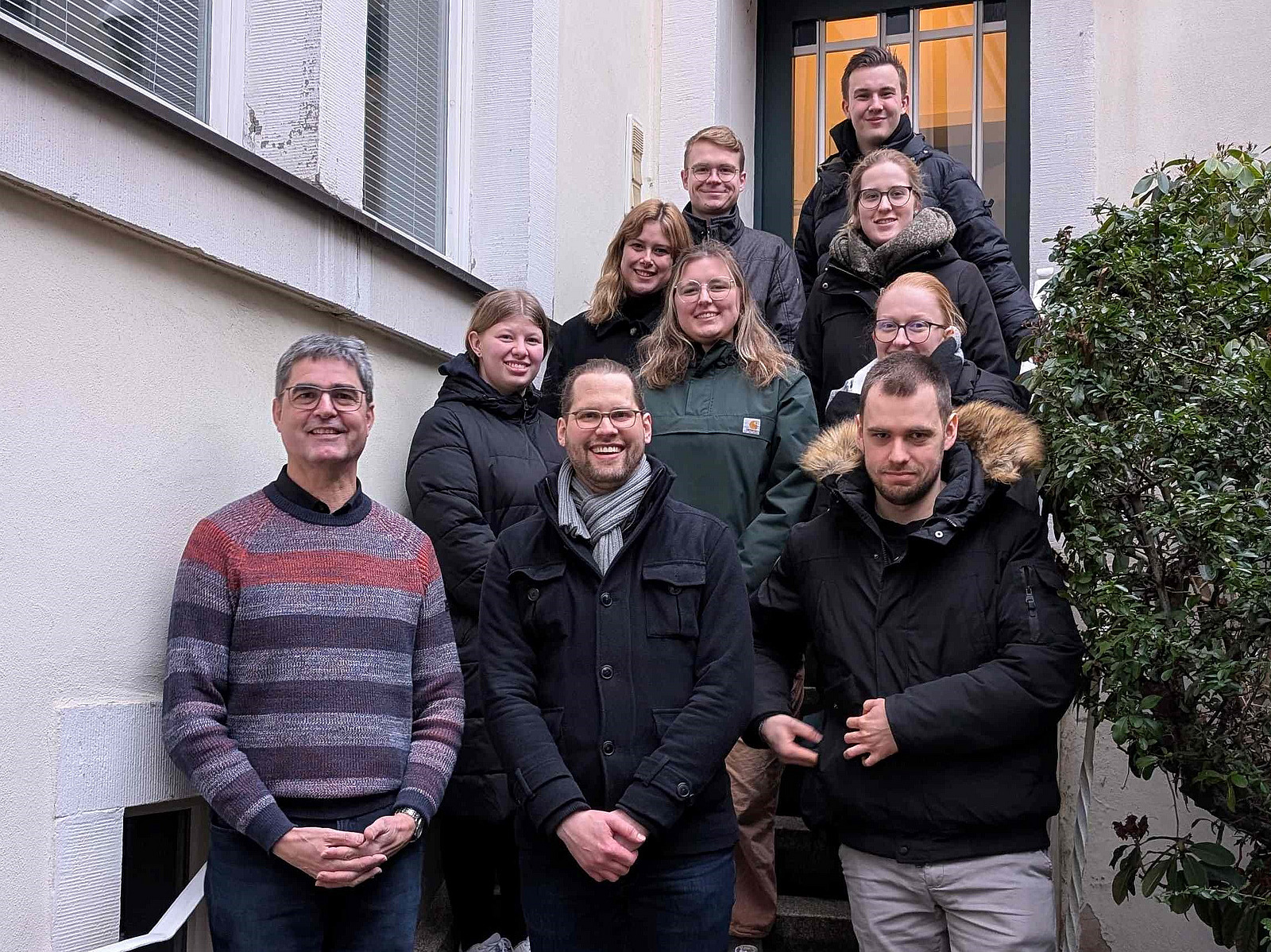 Mehrere Personen stehen und sitzen auf einer Au&szlig;entreppe zwischen zwei Geb&auml;udew&auml;nden. Die Gruppe ist in mehreren Reihen angeordnet und schaut in Richtung Kamera.  Die Personen tragen &uuml;berwiegend Jacken und warme Kleidung. Rechts ist ein Busch oder Strauch zu sehen.  Die Treppe f&uuml;hrt nach oben zu einer T&uuml;r im Hintergrund.