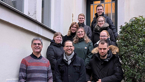 Mehrere Personen stehen und sitzen auf einer Au&szlig;entreppe zwischen zwei Geb&auml;udew&auml;nden. Die Gruppe ist in mehreren Reihen angeordnet und schaut in Richtung Kamera.  Die Personen tragen &uuml;berwiegend Jacken und warme Kleidung. Rechts ist ein Busch oder Strauch zu sehen.  Die Treppe f&uuml;hrt nach oben zu einer T&uuml;r im Hintergrund.