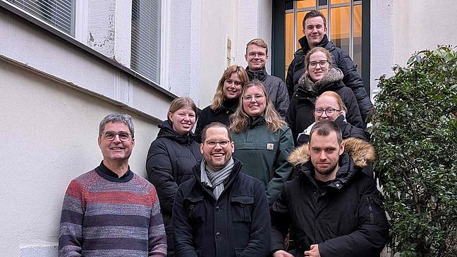 Mehrere Personen stehen und sitzen auf einer Au&szlig;entreppe zwischen zwei Geb&auml;udew&auml;nden. Die Gruppe ist in mehreren Reihen angeordnet und schaut in Richtung Kamera.  Die Personen tragen &uuml;berwiegend Jacken und warme Kleidung. Rechts ist ein Busch oder Strauch zu sehen.  Die Treppe f&uuml;hrt nach oben zu einer T&uuml;r im Hintergrund.