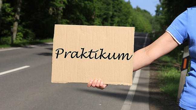Eine Person h&auml;lt ein Schild, auf dem Praktikum steht, im Hintergrund ist eine Stra&szlig;e zu sehen.