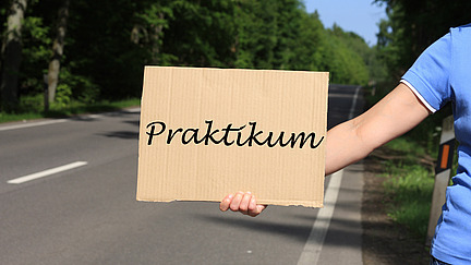 Eine Person h&auml;lt ein Schild, auf dem Praktikum steht, im Hintergrund ist eine Stra&szlig;e zu sehen.