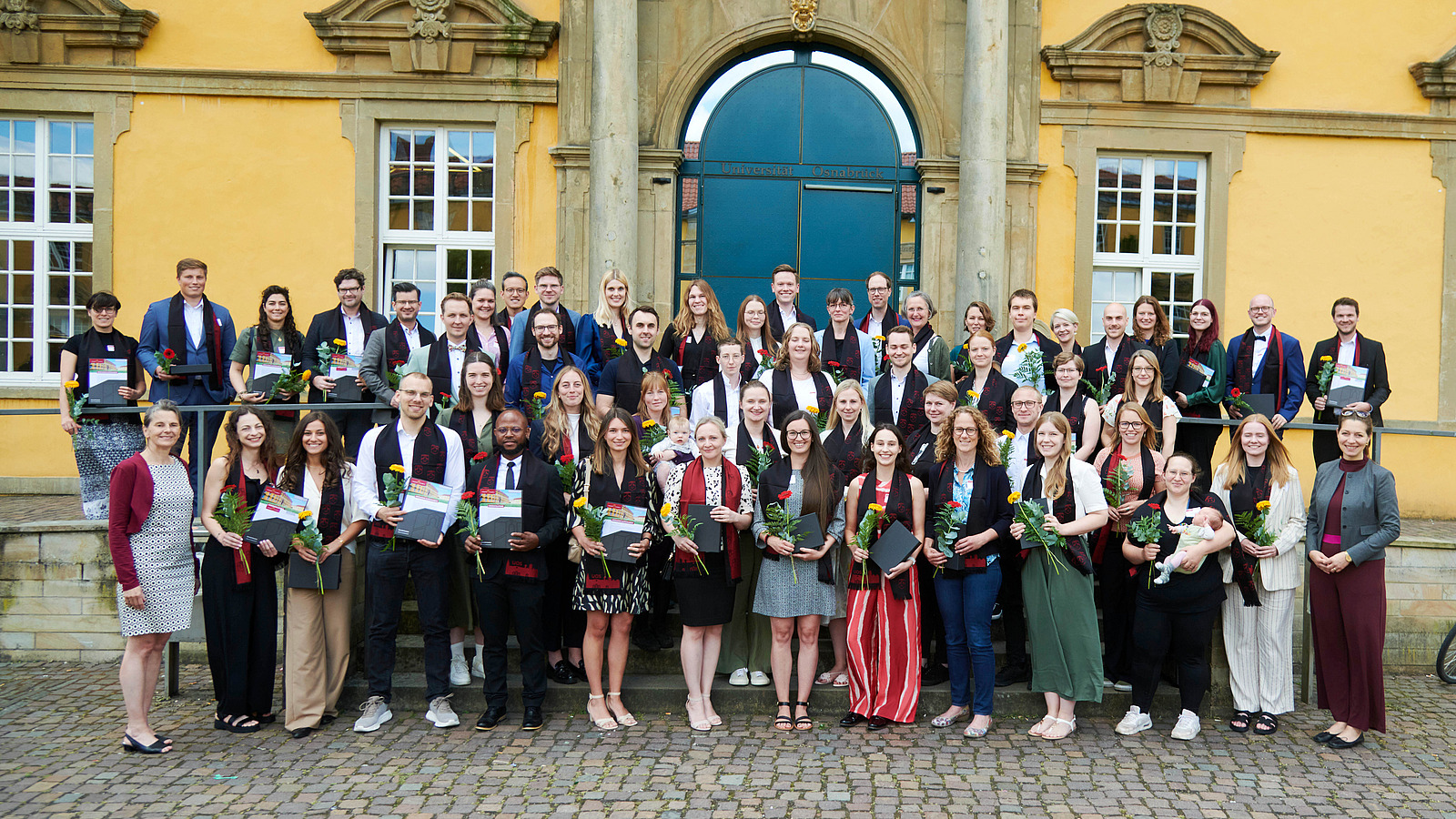 A group of festively dressed people stand in front of a historic building. They are all wearing an elegant scarf and holding a flower and a folder in their hands.