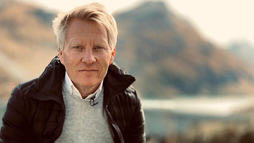 Portraitfoto eines grauhaarigen Mannes vor einer bergigen Landschaft.