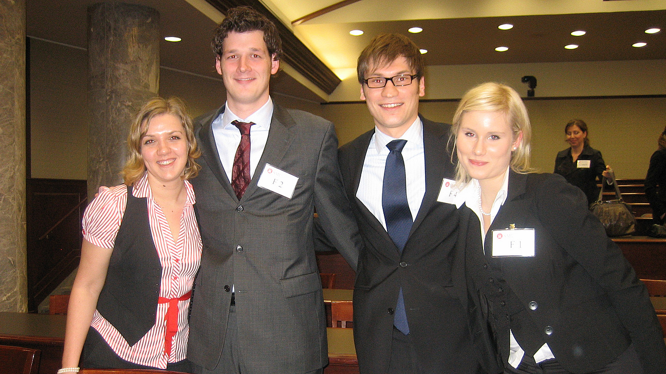 Zwei Frauen und zwei M&auml;nner, f&ouml;hlich l&auml;chelnd, in einer Reihe in einem Seminarraum stehend