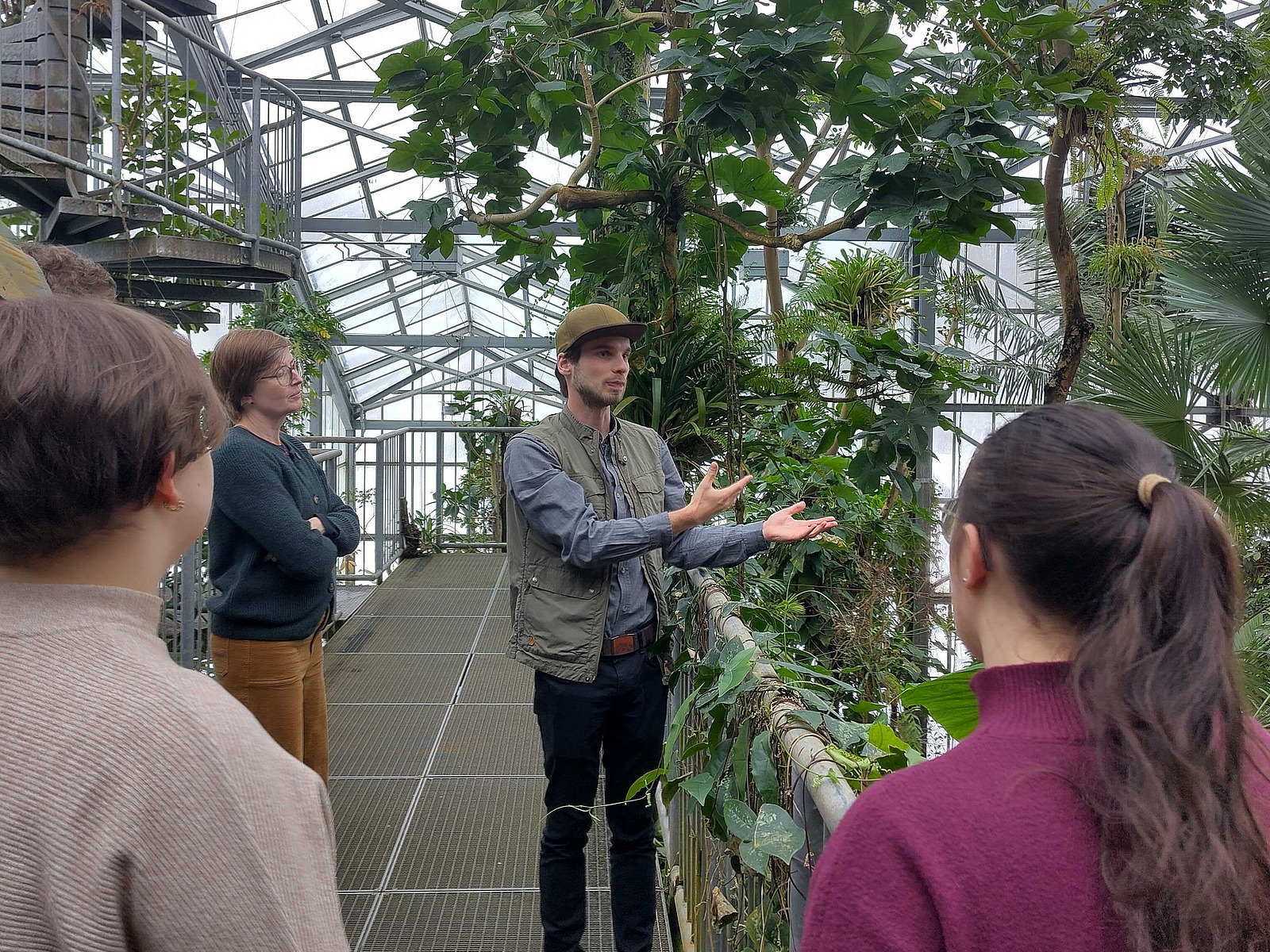 A group of people are standing in a greenhouse filled with green plants, listening to a man who appears to be telling them something.