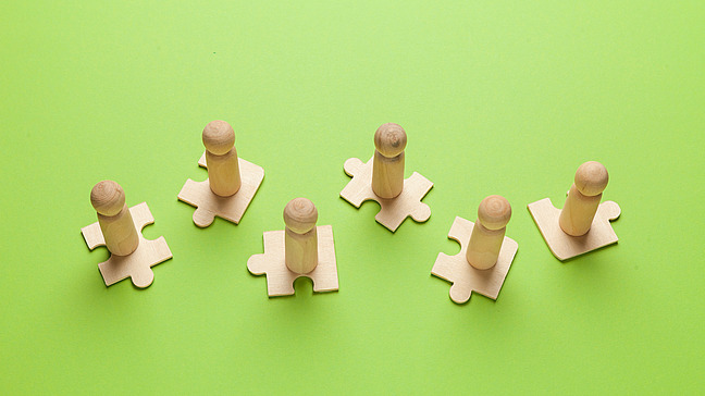 Holzfiguren stehen auf sechs unverbundenen Puzzelteilen.
