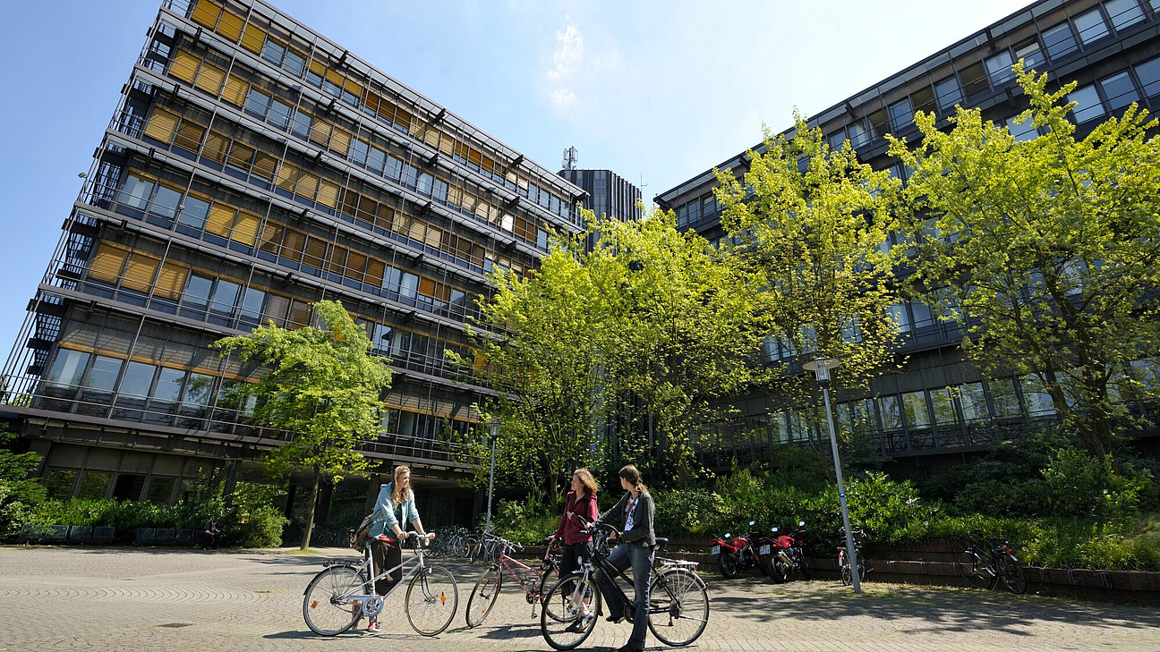 Drei Studierende auf Fahrr&auml;dern vor einem grauen hohen Geb&auml;ude