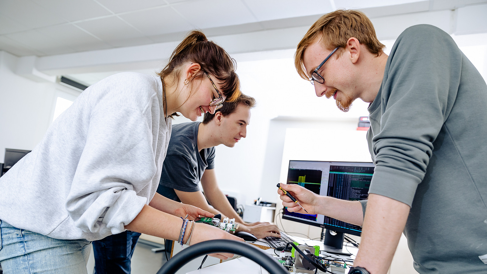 Drei Studierende arbeiten konzentriert an einem technischen Projekt. Sie untersuchen eine elektronische Schaltung und scheinen gemeinsam eine Lösung zu entwickeln.