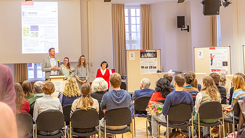 Viele Personen sitzen auf Stühlen und hören einem Vortragenden zu
