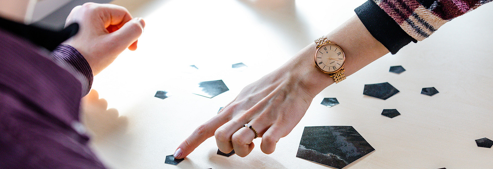 Zwei Personen arbeiten an einem Tisch, auf dem verschiedene geometrische Formen aus schwarzem Material verteilt sind. Eine Person zeigt mit ihrem Finger auf eine der Formen, während die andere Person, auf die Form schaut. Das Licht fällt sanft auf den Tisch, wodurch eine helle Atmosphäre entsteht.
