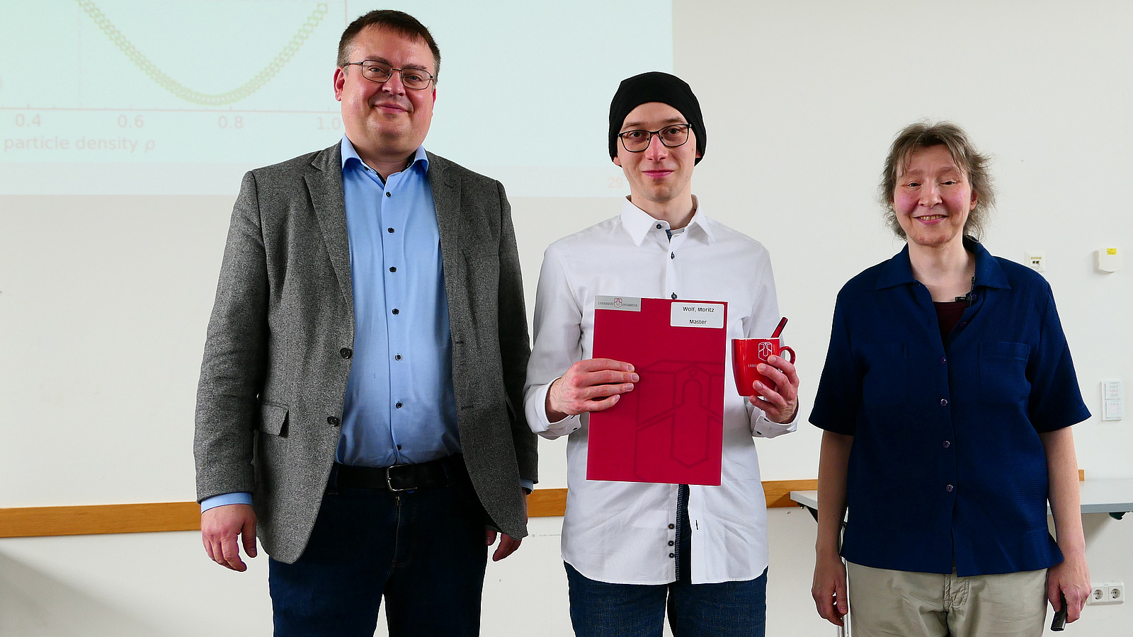 Ein Student mit Studiendekanin und Dekan stehen nebeneinander. Der Student hat eine Mappe in der Hand