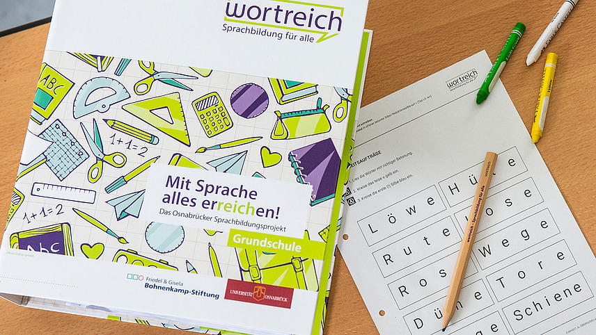 Auf einem Schultisch liegt wortreich Arbeitsmaterial. Neben einem Ordner liegt ein Arbeitsblatt, auf dem man die Wörter Löwe, Rute, Wege oder Schienen erkennen kann.