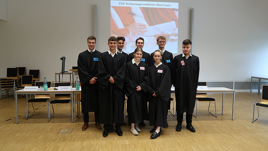 Stehende Gruppe aus 6 M&auml;nnern und 2 Frauen in schwarzen Roben in nachgestelltem Gerichtssaal