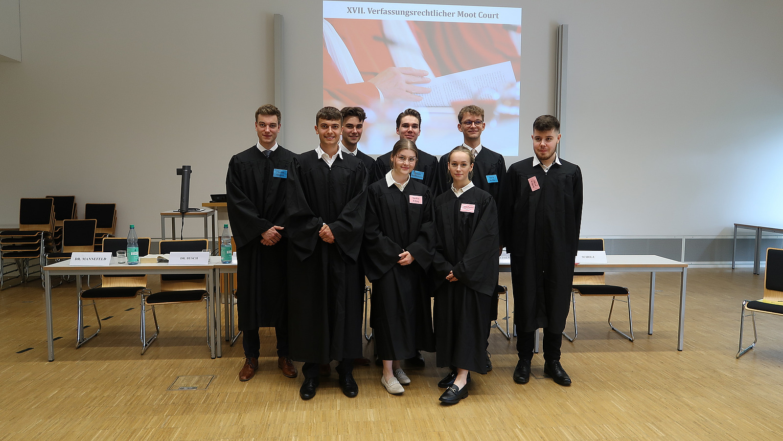 Stehende Gruppe aus 6 M&auml;nnern und 2 Frauen in schwarzen Roben in nachgestelltem Gerichtssaal