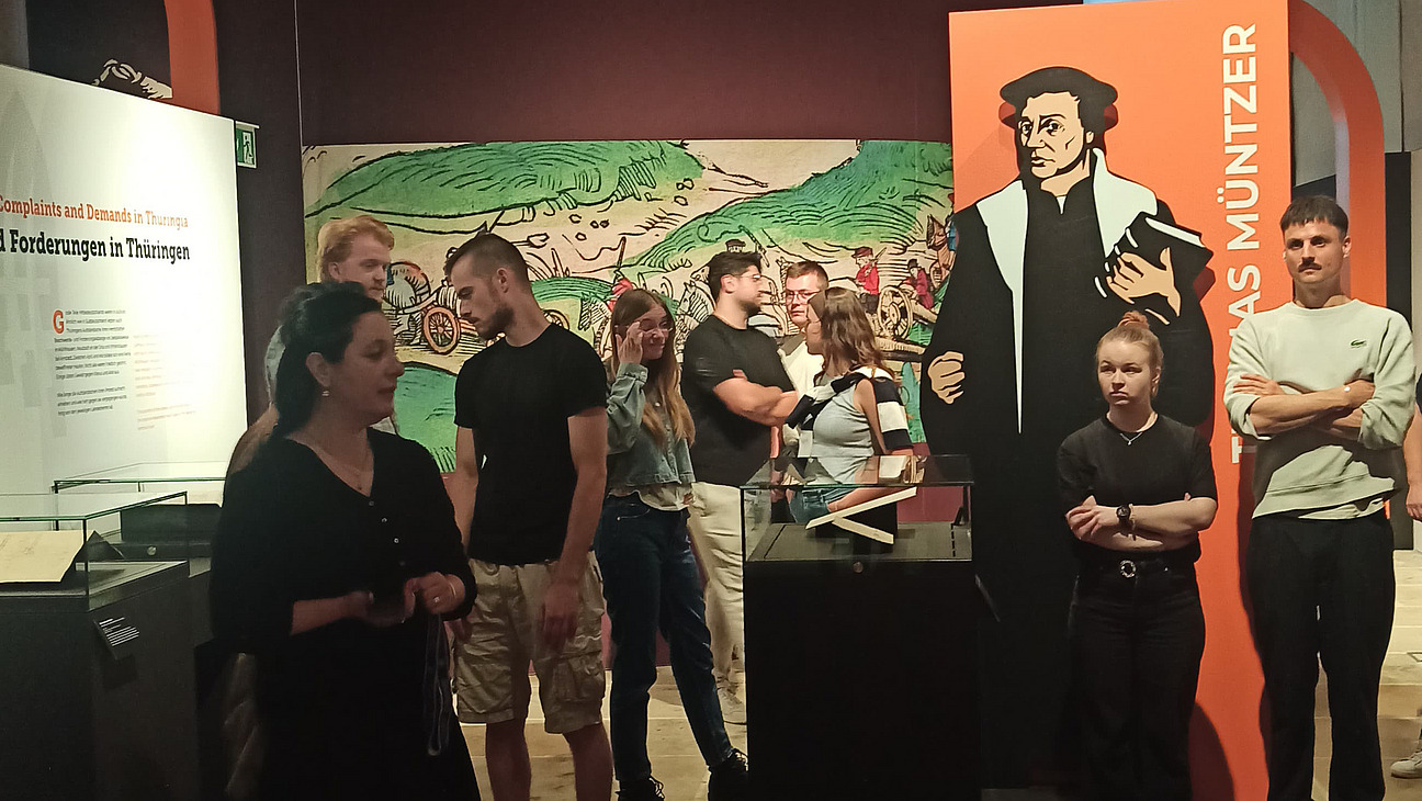 Das Bild zeigt eine Gruppe Menschen, die in einer Ausstellung sind. Die Wände sind orange und schwarz.