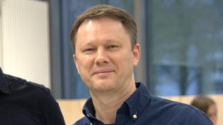 Ein Mann mit kurzen, braunen Haaren und einem freundlichen Gesichtsausdruck steht in einem Innenraum. Er trägt ein dunkelblaues Hemd. Der Hintergrund ist unscharf, zeigt aber Fenster und eine neutrale Einrichtung.