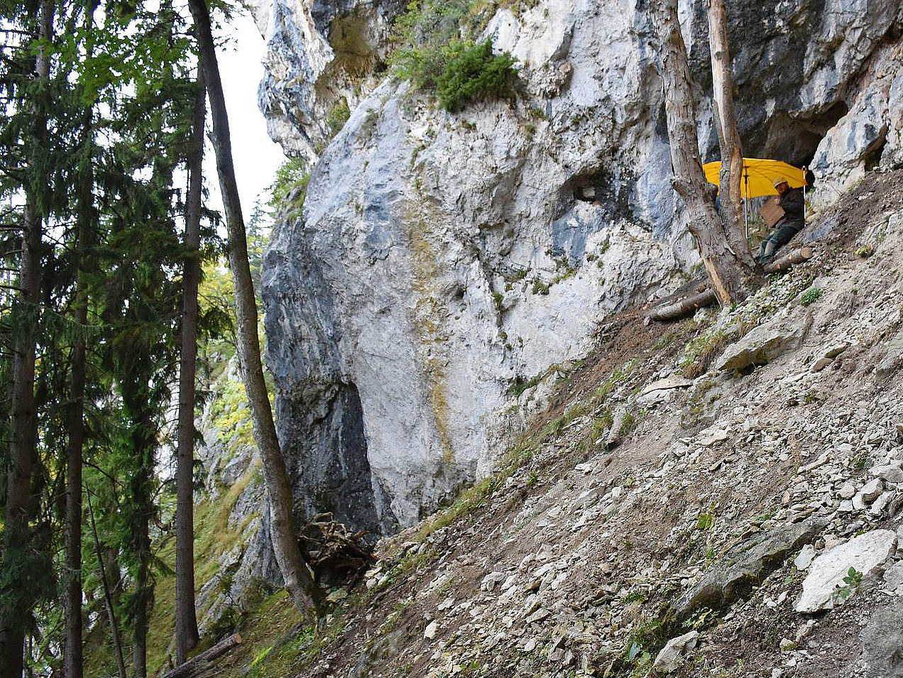 View of a rock face. A person in work clothes and a safety helmet is sitting on the slope in front of the entrance to a cave.