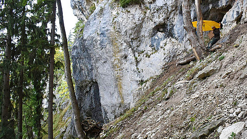 Blick auf eine Felswand. Am Hang sitzt eine Person in Arbeitskleidung und Sicherheitshelm vor dem Eingang einer Höhle