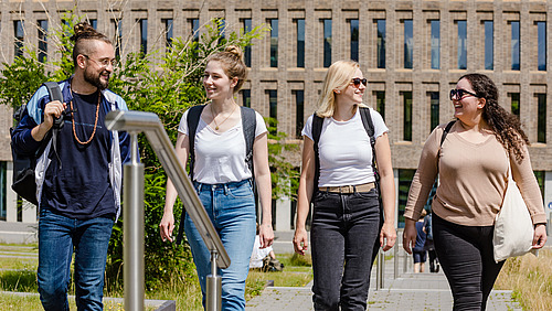 Vier Studierende unterhalten sich beim gehen auf dem Campus, im Hintergrund ist die Biliothek am Westerberg zu sehen