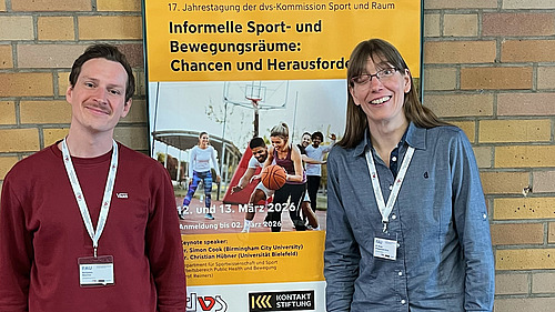 Dr. des. Benjamin B&uuml;scher und Dr. Nicole Br&uuml;ggemann-Kons stehen vor einem Poster zum informellen Sport.