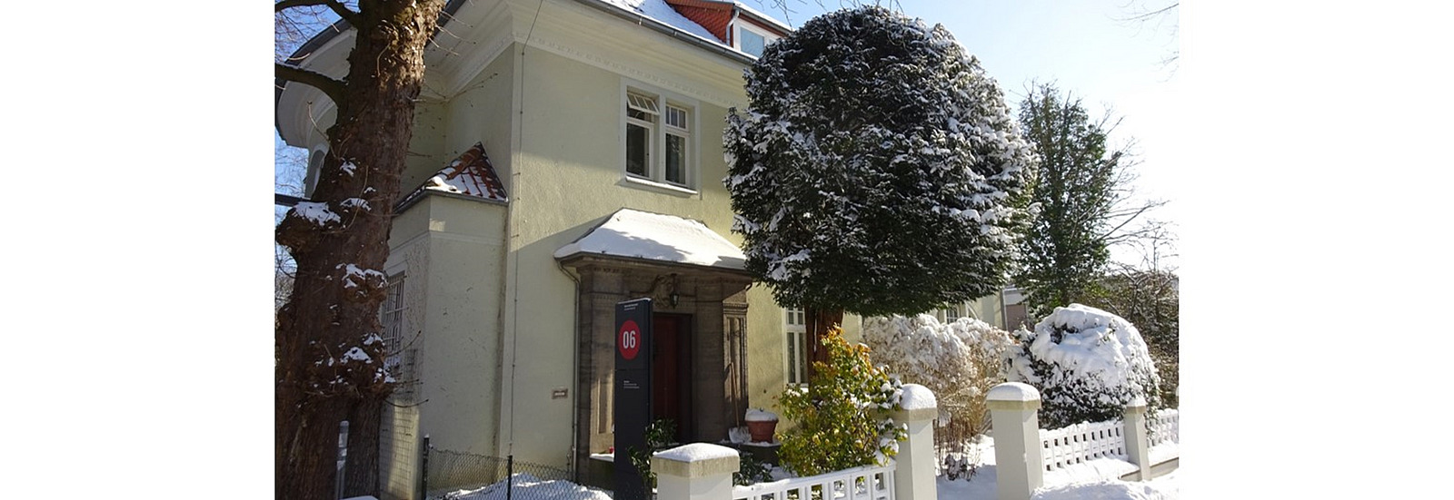 Street view of the guest house in winter, with snow on the roof, the trees and bushes and on the railings.