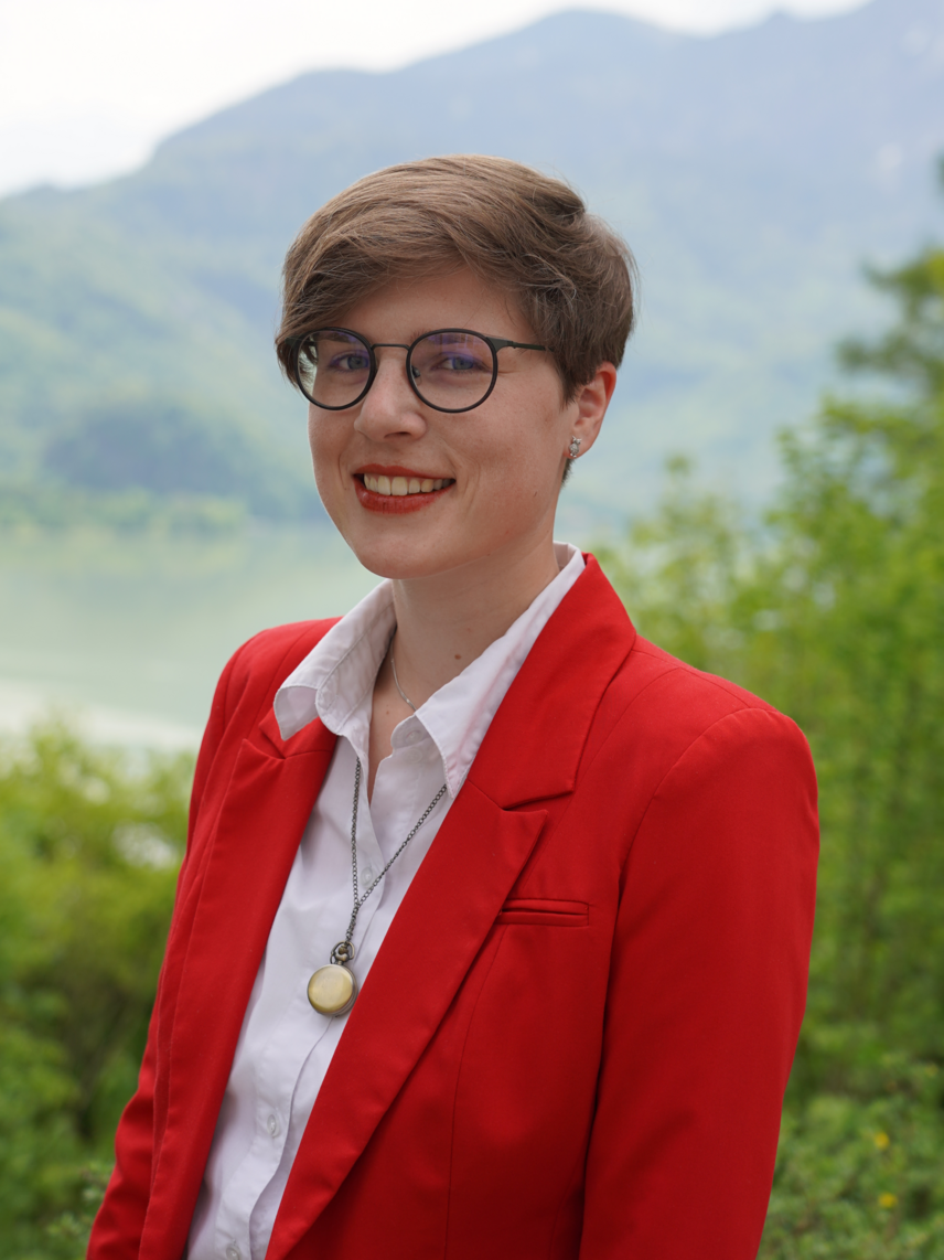   Eine Frau mit Brille und kurzen Haaren trägt einen roten Blazer und ein weißes Hemd. Um den Hals trägt sie ein Medaillion an einer Kette. Sie steht vor einer Kulisse aus grünen Bäumen und einem verschwommenen Berg im Hintergrund. 