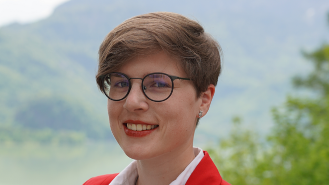   Eine Frau mit Brille und kurzen Haaren trägt einen roten Blazer und ein weißes Hemd. Um den Hals trägt sie ein Medaillion an einer Kette. Sie steht vor einer Kulisse aus grünen Bäumen und einem verschwommenen Berg im Hintergrund. 