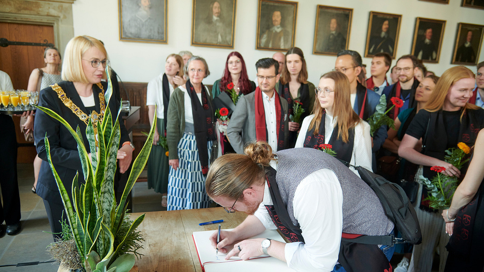 In a historic-looking building, several people are standing at a table on which an open book with blank pages is lying. One person is writing something in the book.
