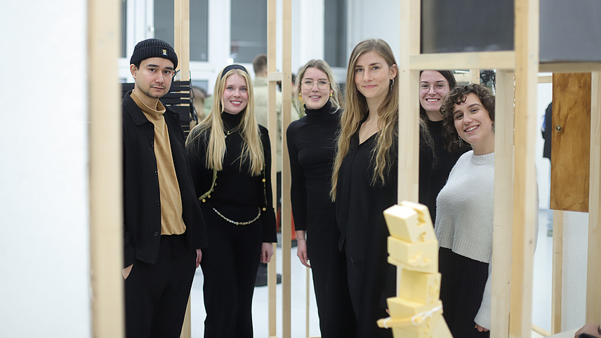 Six students, dressed mostly in black, are standing in an art exhibition and smiling at the camera.