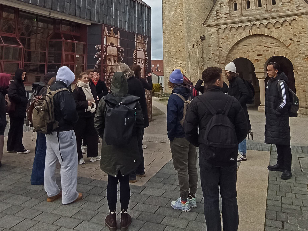 Eine Gruppe steht auf einem Platz. Links ist ein graues, mit Metal verkleidetes Gebäude; im Hintergrund eine helle Kirche mit grünem Dach und einem Turm. 