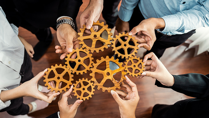 A group of elegantly dressed people from different countries hold wooden cogwheel parts in their hands. The close-up of the cogwheels held together shows their interaction.