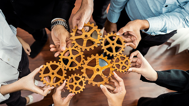 A group of elegantly dressed people from different countries hold wooden cogwheel parts in their hands. The close-up of the cogwheels held together shows their interaction.