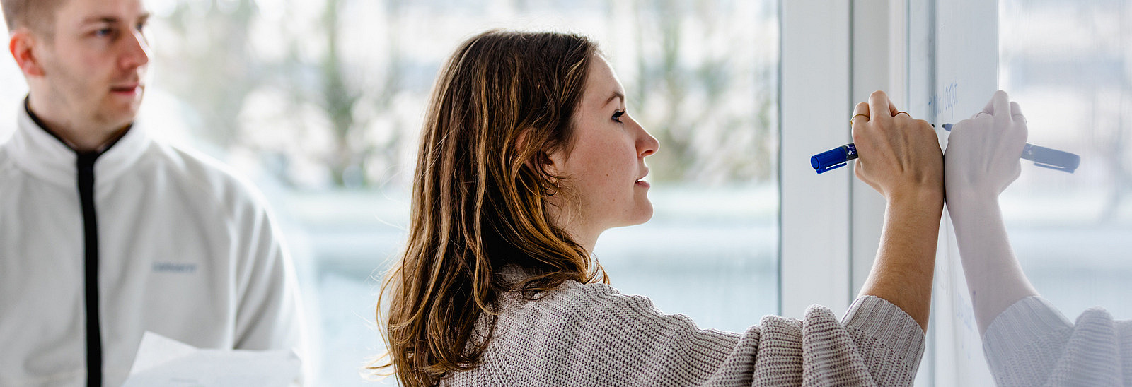 Eine Frau schreibt mit einem blauen Marker auf ein Whiteboard. Sie hat braune Haare und trägt einen grauen, weiten Pullover. Im Hintergrund steht ein männlicher Kollege in einem weißen Oberteil und hält eine gedruckte Seite in der Hand, mit Blick auf das Whiteboard. Durch die Fenster ist natürliches Licht zu sehen, das den Raum hell erleuchtet, während die Außenansicht leicht verschwommen bleibt.