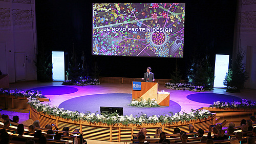 Ein Mann steht auf einem Podium an einem Rednerpult, im Hintergrund ist der Schriftzug "De Novo Protein Design" zu sehen.