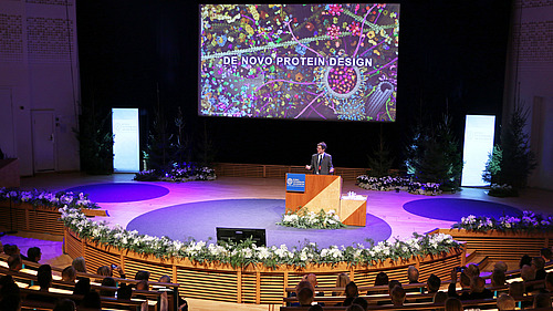 Ein Mann steht auf einem Podium an einem Rednerpult, im Hintergrund ist der Schriftzug "De Novo Protein Design" zu sehen.