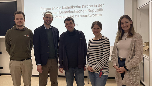F&uuml;nf Personen stehen nebeneinander in einem Raum und schauen in Richtung Kamera.  Hinter ihnen ist eine helle Wand mit projiziertem Text zu sehen. Der Text beginnt mit &bdquo;Fragen an die katholische Kirche in der Deutschen Demokratischen Republik &hellip;&ldquo;.  Die Personen stehen in einer Reihe und nehmen den Vordergrund des Bildes ein.