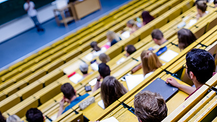 Blick von oben auf Sitzreihen in einem H&ouml;rsaal. Die Studierenden machen sich Notizen auf ihren Bl&ouml;cken.