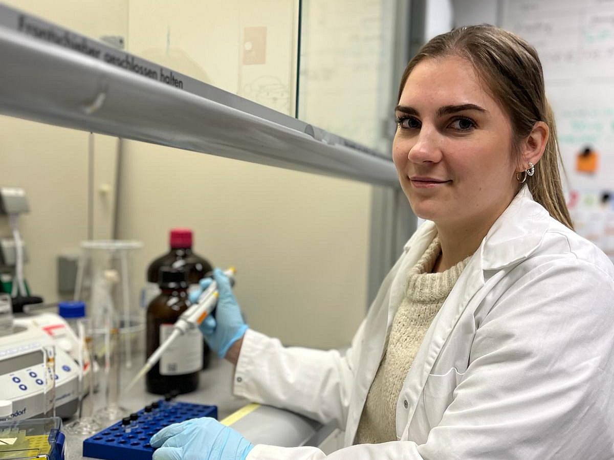 A woman wearing a white coat is sitting at a laboratory bench holding a pipette.
