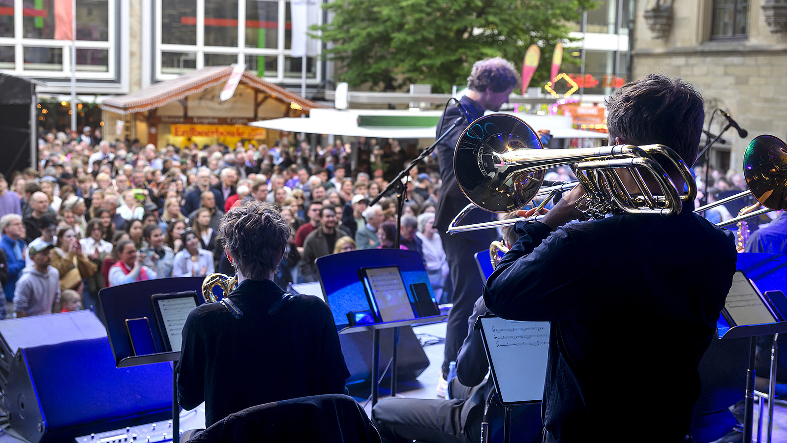 Several brass players from behind on a stage, with spectators in the background