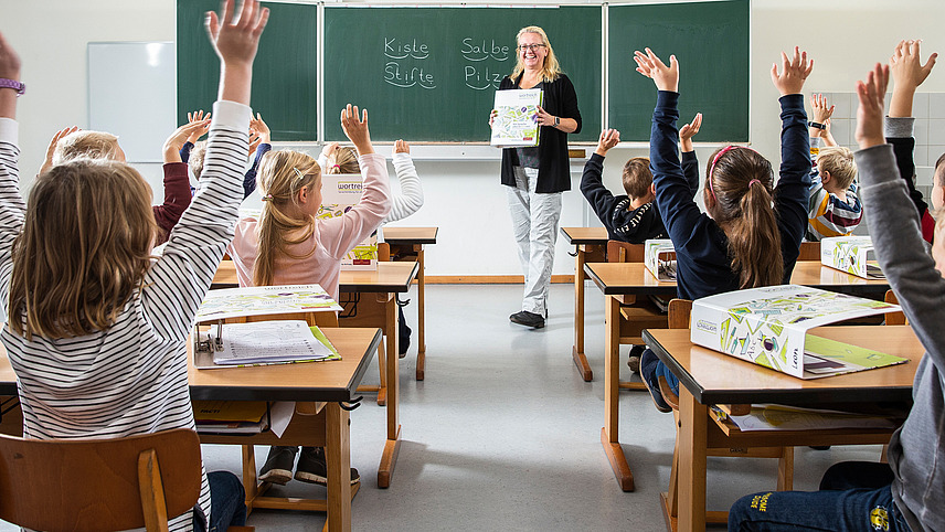 Eine Lehrerin steht vor einer Tafel und zeigt einer Grundschulklasse einen Wortreich-Ordner. Die Schüler und Schülerinnen halten einen Arm in die Höhe, um sich zu melden.