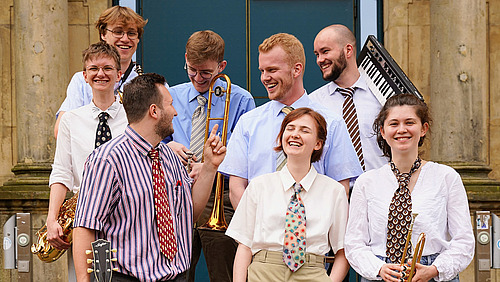A group of musicians stands in front of a historic building, smiling at the camera. The musicians are playing various instruments, including saxophone, trombone, and keyboard.